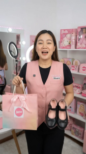 Barbie-themed black Mary Jane shoes with pink accents and decorative bow. Pink gradient gift bag with Barbie logo and ribbon. Store employee in pink vest holding items.