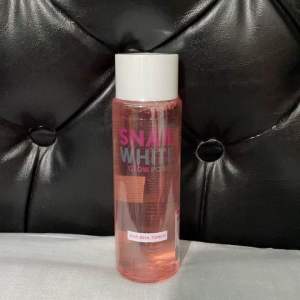SNAIL WHITE GLOW AHA-BHA Toner 150ml - Clear bottle with pink liquid white cap label on front held in hand against black tufted background.