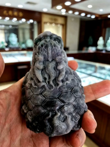 Myanmar jade longgui with black green and purple colors. Detailed carving smooth texture intricate design natural stone decorative item. Displayed in a jewelry store setting.