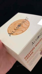 Kem dưỡng trắng da Almonds Prestige Whitening Cream dung tích 490K thiết kế lọ thủy tinh màu nâu đặt trên bề mặt cong màu be xung quanh có các viên đá nhỏ trong suốt.