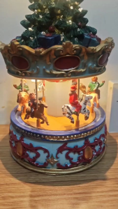Christmas carousel music box with rotating horses and Santa figure featuring a decorated tree and star on top ornate blue and red base and mounted on a wooden surface.