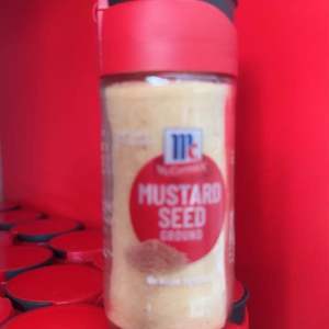 McCormick Mustard Seed Ground 26g Small Bottler Cooking & Seasoning - Red cap clear container visible mustard seeds red label with brand logo placed on red shelf.