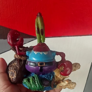 Purple crab figurine with green eyes and red claws holding a brown sack with gold design. Wearing blue hat and belt. Scene: aquarium decoration.