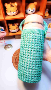 Túi đựng bình giữ nhiệt size 1500ml màu xanh lá cây đan móc có quai xách đặt trên đế mây trong không gian trang trí với kệ gỗ và thú nhồi bông.