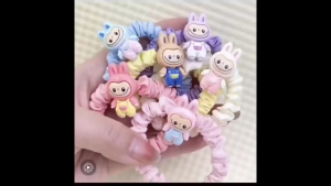 Pastel-colored hair scrunchies with decorative charms in assorted colors including pink purple yellow and white. Each scrunchie is packaged in a clear plastic bag.