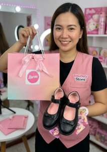 Barbie-themed black Mary Jane shoes with pink accents and decorative bow. Pink gradient gift bag with Barbie logo and ribbon. Store employee in pink vest holding items.