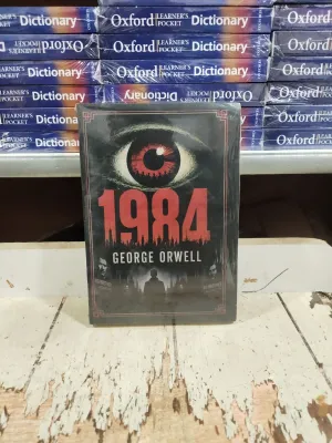 Buku 1984 karya George Orwell dengan sampul hitam dan mata merah di atas meja kayu dikelilingi kamus Oxford di latar belakang.