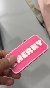 Pink rectangular name tags with white raised letters. One tag reads "JENNY" and the other "CHERRY". Both have a hole for attachment.