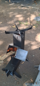 Heavy Duty Modern Rocket stove Wood Burning Grill-stove (Kalan De Kahoy)MakapaL Outdoor metal fire pit with hopper and ash catcher on grass near stone wall.Outdoor Metal Grill Stands with Fuel Hopper and Cooking Surface.
