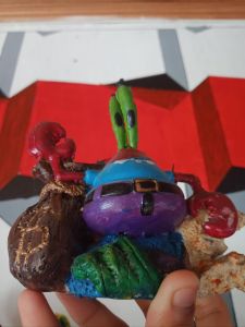 Purple crab figurine with green eyes and red claws holding a brown sack with gold design. Wearing blue hat and belt. Scene: aquarium decoration.