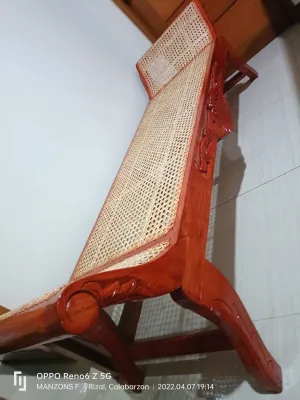 Cleopatra Bench (Wooden lounge chair with woven cane seat and backrest. Ornate carvings on frame. Suitable for indoor relaxation. Positioned against a tiled wall.)