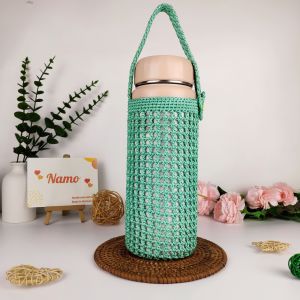 Túi đựng bình giữ nhiệt size 1500ml màu xanh lá cây đan móc có quai xách đặt trên đế mây trong không gian trang trí với kệ gỗ và thú nhồi bông.