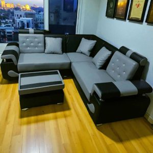 Sofa ruang tamu model L angsa terbaru BJ08 dengan bantalan abu-abu dan hitam dilengkapi meja kopi kecil di ruangan dengan lantai kayu dan pemandangan kota di malam hari.