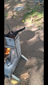 Heavy Duty Modern Rocket Wood Burning Grill (The TOWER-Kalan De Kahoy) MakapaL Outdoor metal fire pit with hopper and ash catcher on grass near stone wall.Outdoor Metal Grill Stands with Fuel Hopper Outdoor black metal stove with open door and visib