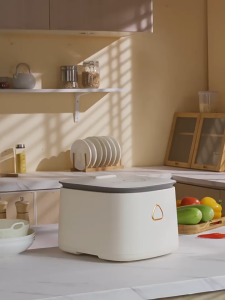 White 3L Multi-Function Rice Cooker with digital display and triangular logo. Suitable for steaming and cooking rice. Set on wooden table with rice bowl and cooked dishes.