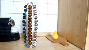 Stainless steel coffee capsule holder with multiple colored capsules next to coffee machine and cup.
