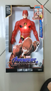The Flash action figure with yellow lightning bolt emblem and red and yellow accents standing upright on a white surface with a small red cylindrical object beside it.