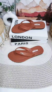 Brown leather flip flops with white stitching and metallic logo on footbed. Placed on white books with "LONDON" text. On textured white rug.