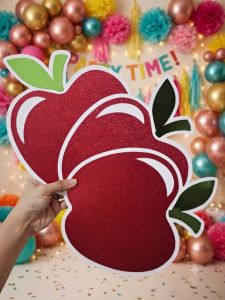 Red glitter apple cutouts held by hand in festive party setting with balloons and decorations.