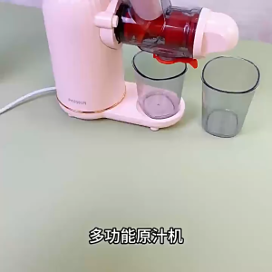 Cream-colored slow cold press juicer with transparent chute and black spout. Juice and pulp collected in separate containers. Person placing carrot into juicer. Strawberries and carrots on table.
