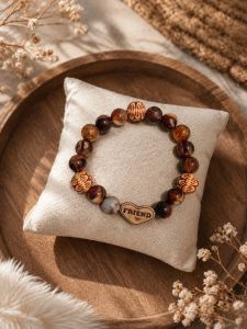 Wooden beaded bracelet with heart charm engraved FRIEND. Natural brown and amber tones. Displayed on cushioned pillow.