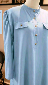 Light blue long tunic with button front and chest pockets. Adjustable sleeves and tailored fit. Suitable for casual or modest wear. Displayed on a mannequin in a home setting.