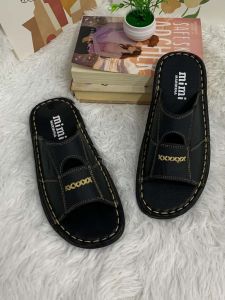 Black slip-on sandals with white stitching and gold lettering on a fluffy white surface.