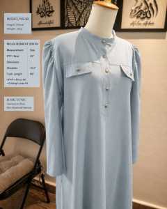 Light blue long tunic with button front and chest pockets. Adjustable sleeves and tailored fit. Suitable for casual or modest wear. Displayed on a mannequin in a home setting.